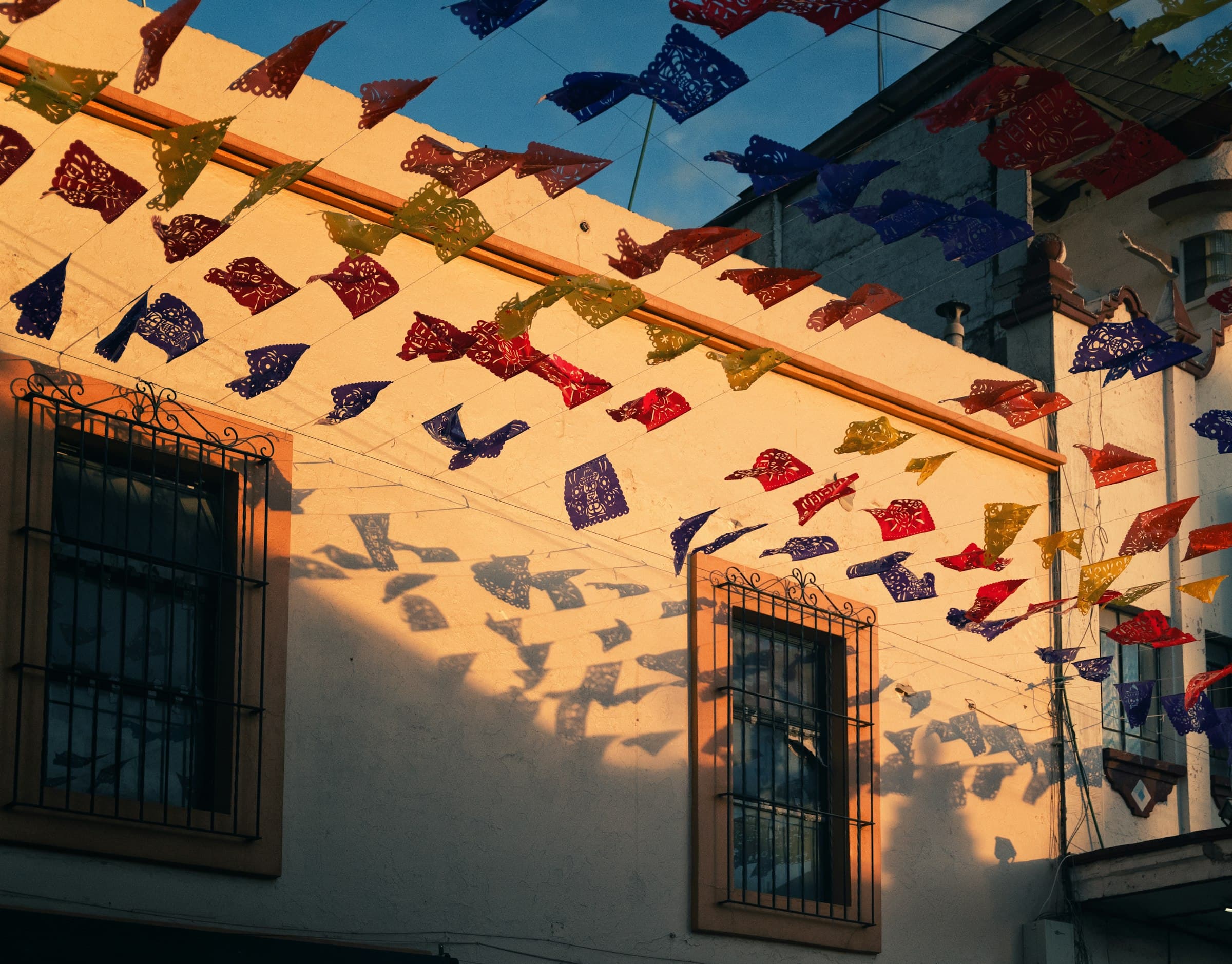 Papel picado de colores colgado sobre una calle, con la luz del atardecer en la fachada de una casa